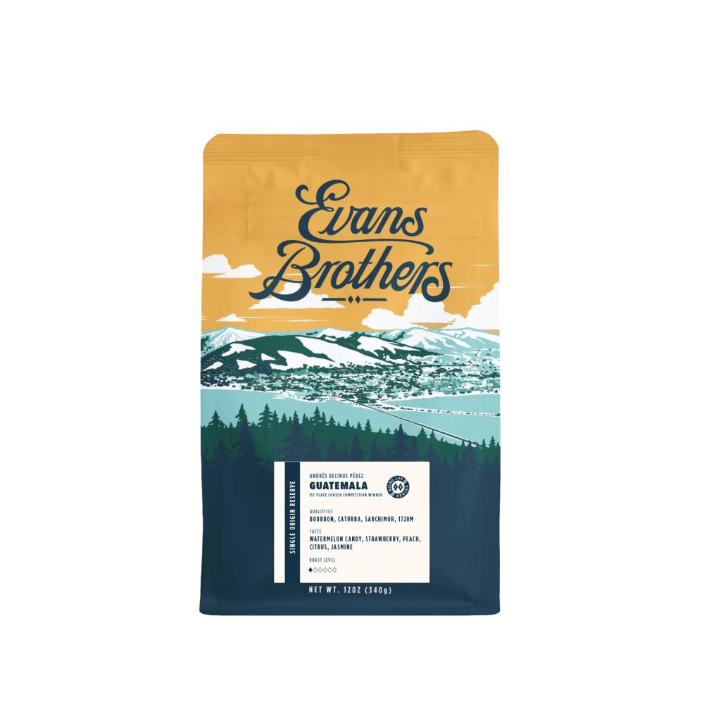 A bag of Evans Brothers coffee from their Guatemala Farm to Table line, featuring a scenic illustration of a mountain town, lake, and pine forest on the front, set against a plain white background. – Evans Brothers Coffee main product